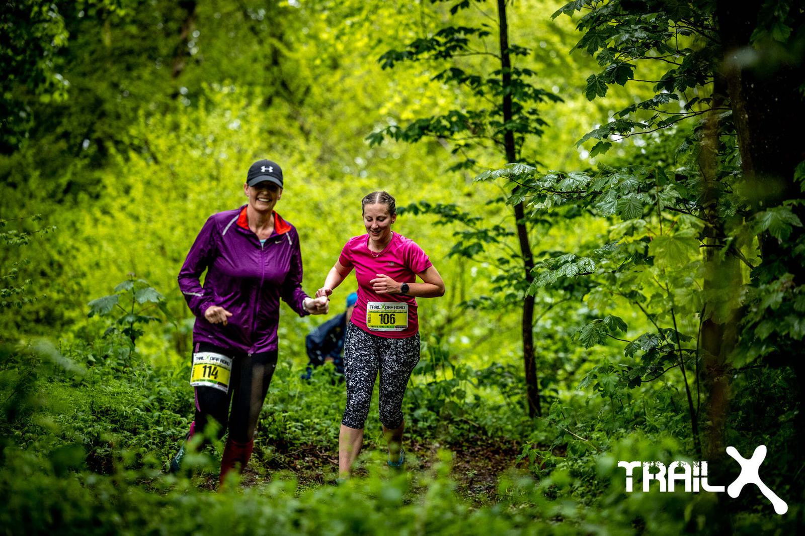 Two women are running on a trail in a forest, wearing race bibs number 114 and 106. They are surrounded by lush green foliage. The woman in the purple jacket is assisting the other wearing a pink top. The word "TRAILX" appears in the bottom right corner.