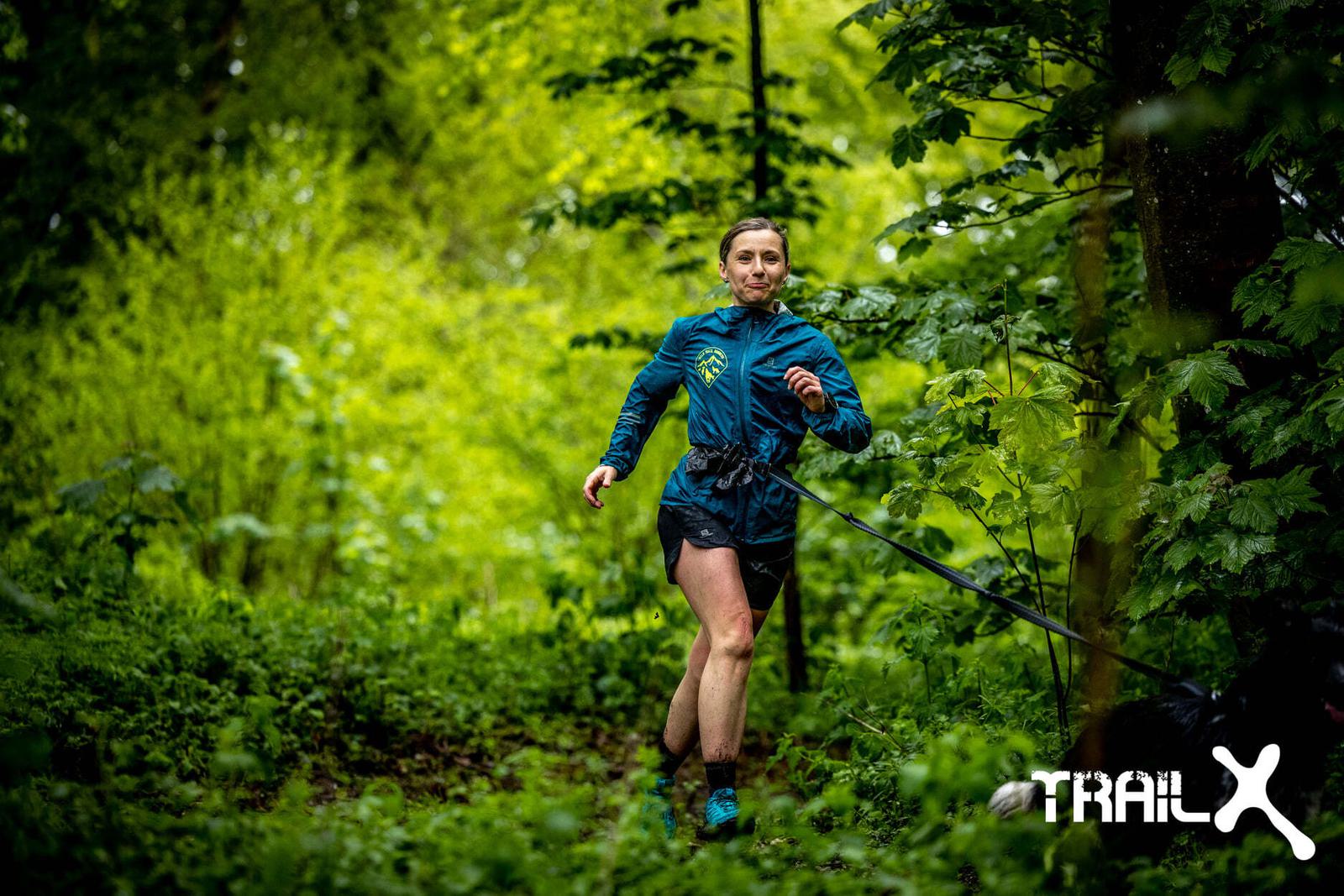 A person wearing a blue jacket and black shorts is running through a lush, green forest trail. Their face shows determination. A black dog, partially visible, appears to be on a leash running beside them. The word "TRAIL" is visible in the bottom right corner.
