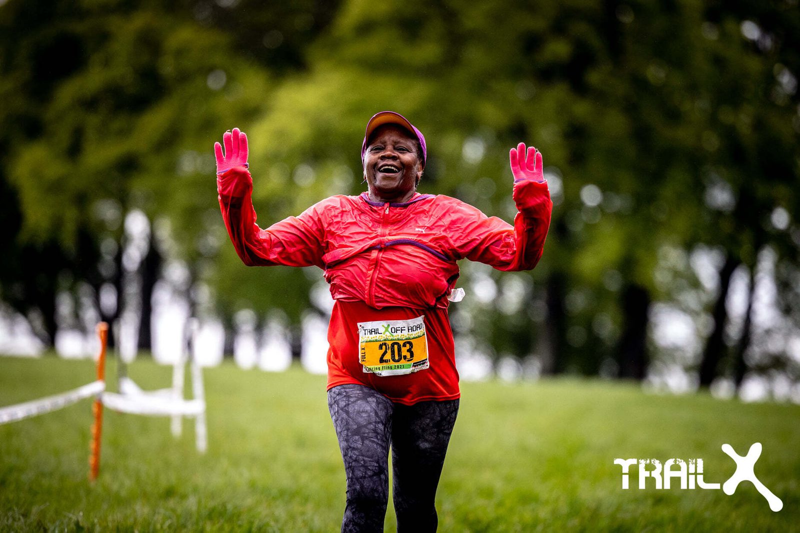 A runner wearing a red jacket and pink gloves smiles and raises her arms while participating in a trail run. Her race bib number is 203. The grassy trail is lined with trees and white tape, and the logo "TRAIL X" is visible in the bottom right corner.