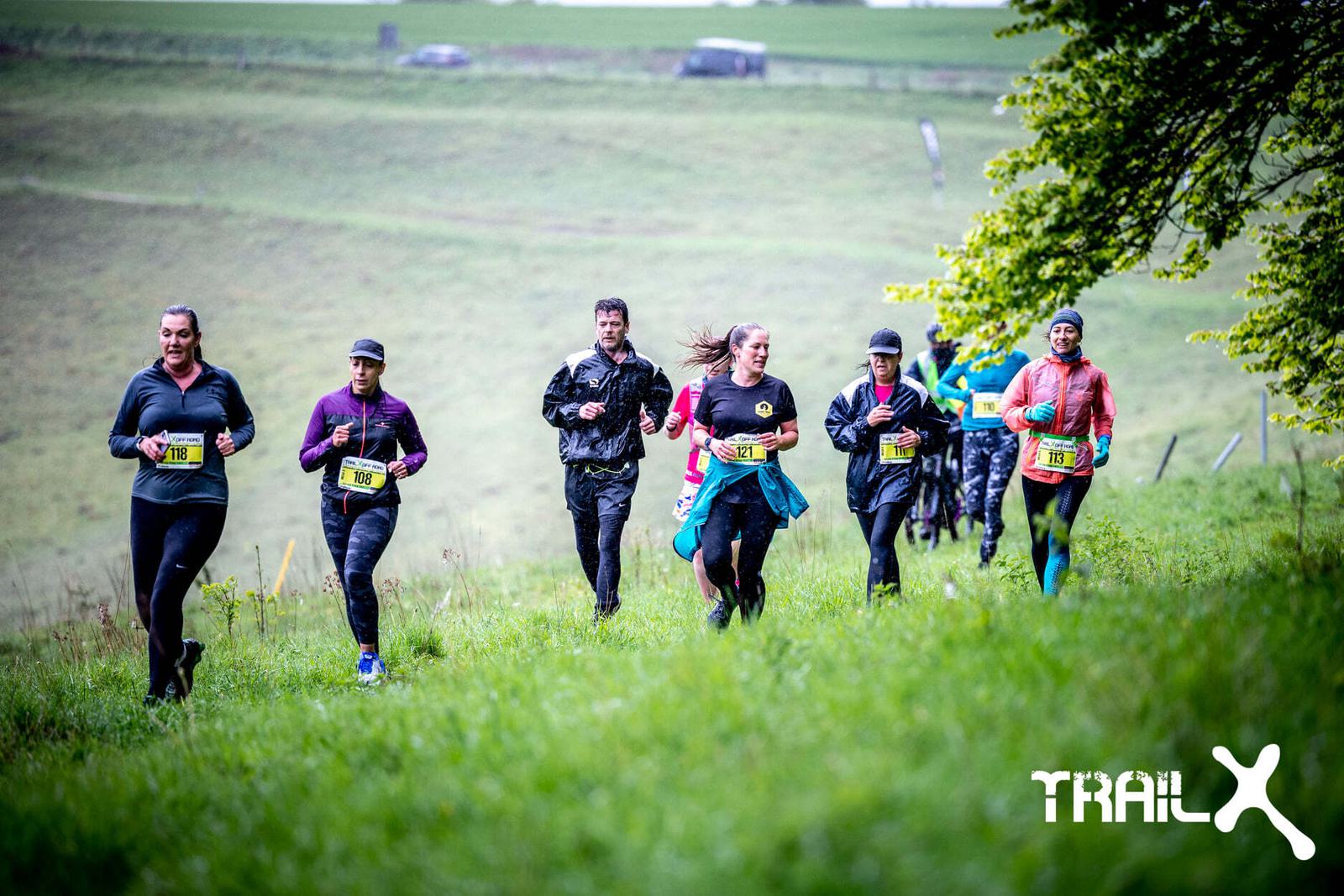 A group of runners in athletic gear are running on a grassy trail. They wear race bibs and appear focused. The landscape is lush and green, with gentle hills in the background. The Trail X logo is visible at the bottom right of the image.