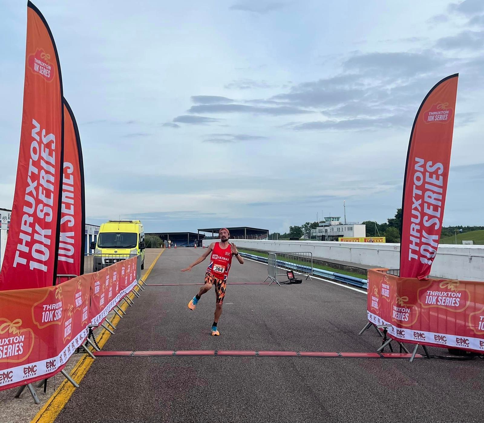 A runner crosses the finish line of the Thruxton 10K Series, surrounded by event banners and a cloudy sky. A yellow van is parked beside the track.