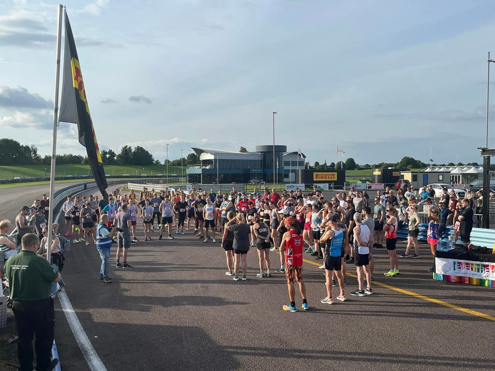 A large group of runners gathers on a racetrack for an event. Participants wear various athletic outfits. A person with a flag stands at the front. The background features buildings and lush greenery under a partly cloudy sky.