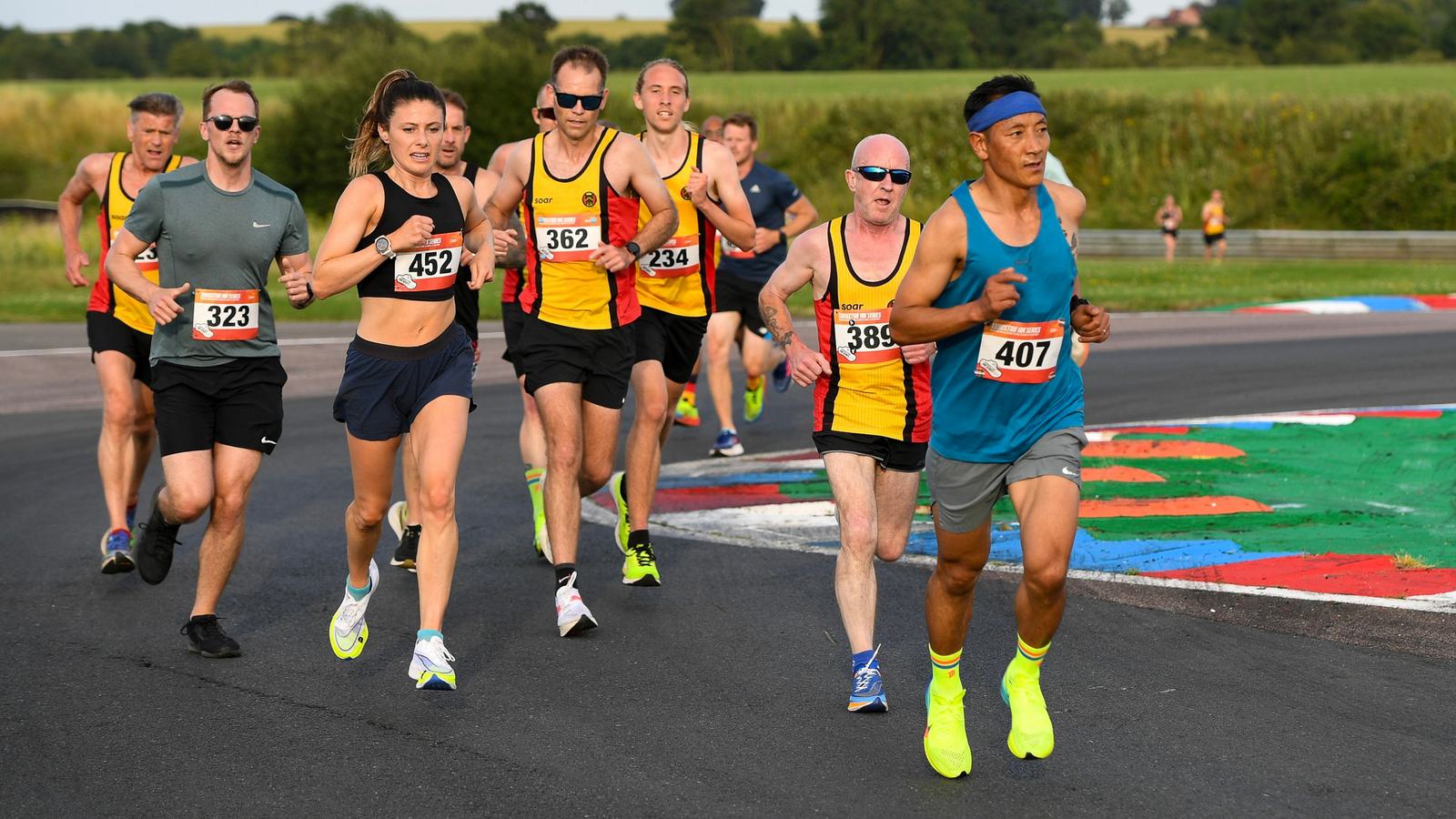 A group of runners competing on a race track. They wear numbered bibs and athletic gear. The background features green fields and trees under a clear sky. One runner in blue leads the pack, with others close behind in yellow tops.