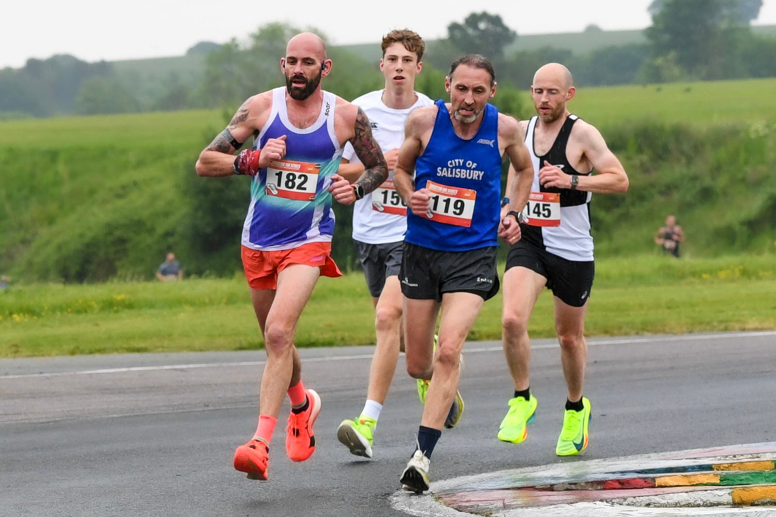 Four men are running a race on a track with a grassy landscape in the background. They wear racing bibs and athletic gear, focusing on speed and competition.