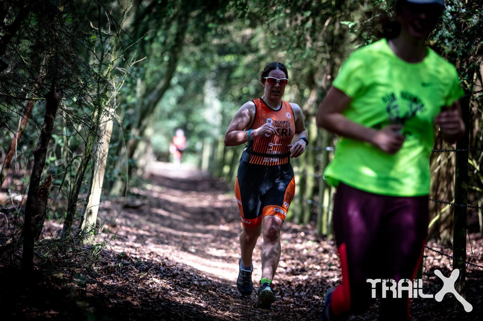 A person wearing a triathlon suit and sunglasses is running on a forest trail. Another runner, wearing a bright green shirt, is partially visible on the right. Sunlight filters through the trees, highlighting the pathway. The word "TRAIL" is visible at the bottom right.