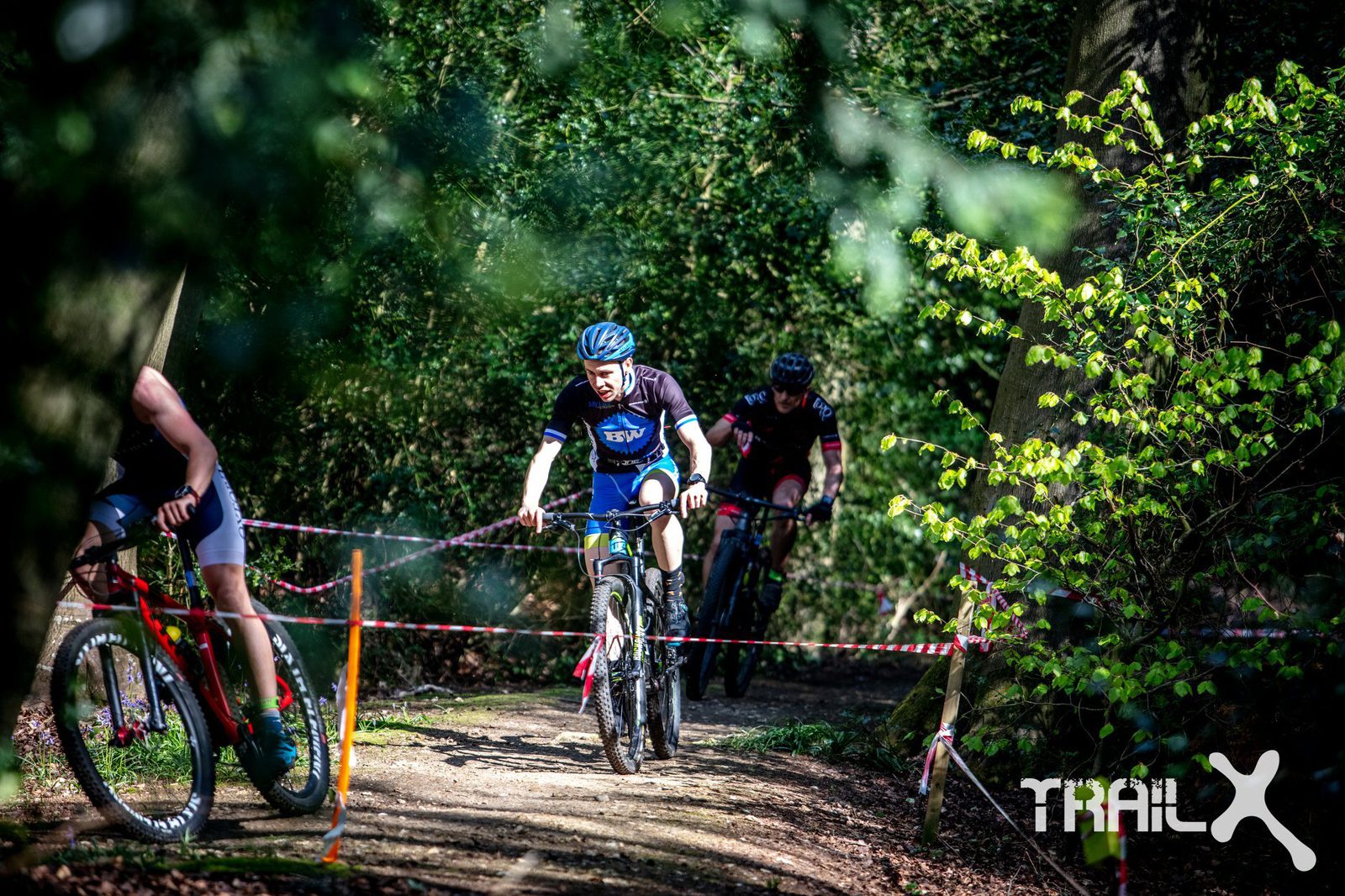 Cyclists ride along a forested trail during a race, marked by red and white tape. The scene is bright with sunlight filtering through the trees. The cyclist in the front appears focused, with others close behind. A banner with the "TRAILX" logo is visible on the right.