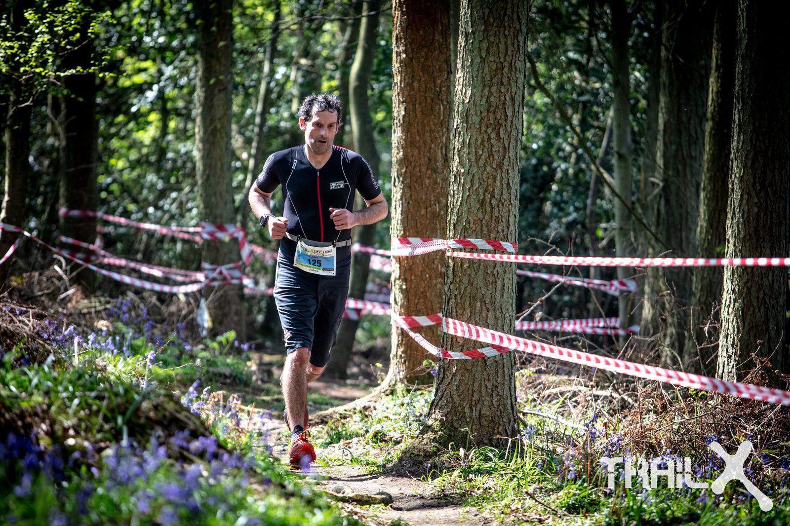 A man in athletic clothing runs on a forested trail, surrounded by trees and red-and-white barrier tape marking the route. He wears a race bib on his chest and bright red running shoes. Purple wildflowers bloom along the side of the trail. The day is sunny.