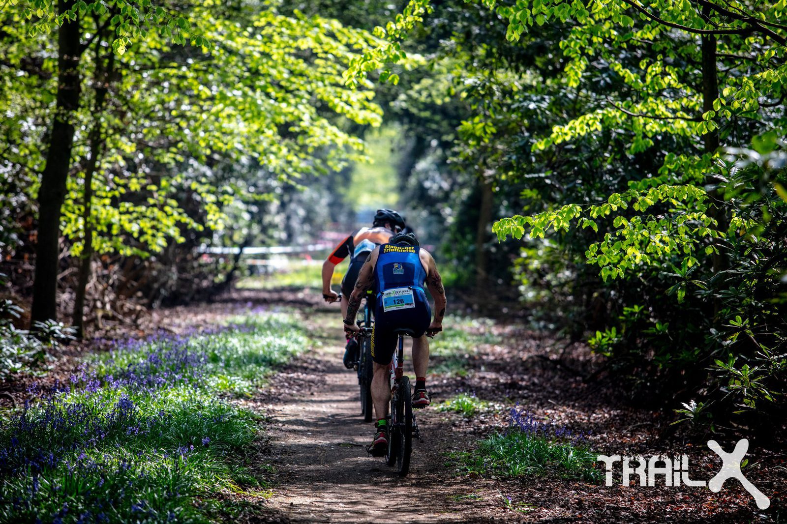 A cyclist wearing a helmet and a numbered bib rides along a dirt trail through a forested area with lush green foliage and patches of blue flowers. The trail is dappled with sunlight filtering through the trees. The word "TRAILX" is visible in the bottom right corner.