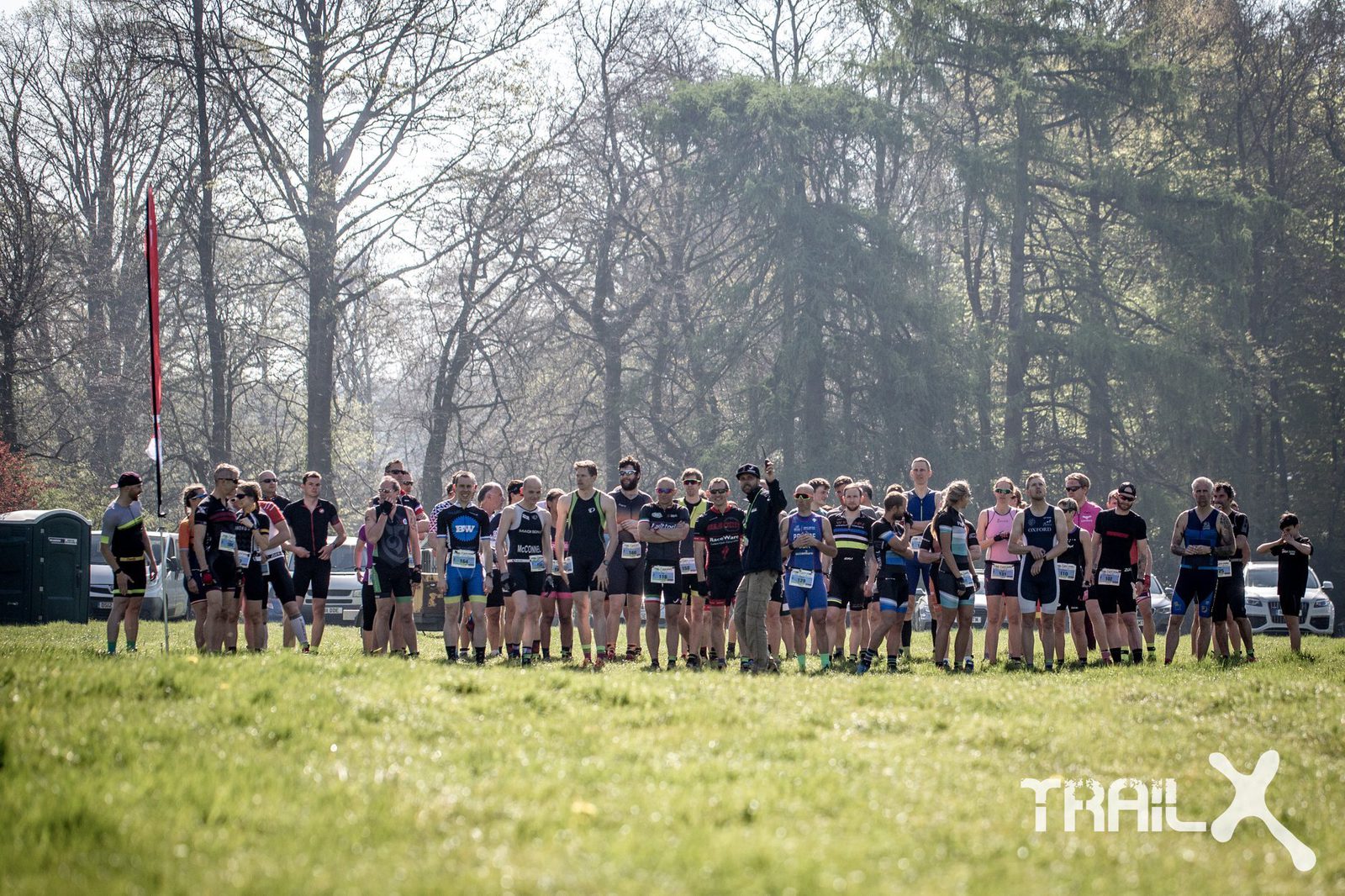 A group of athletes stands at the starting line of a trail running event, wearing race bibs and athletic gear. They are outdoors on a grassy field with trees in the background. A few portable restrooms are visible on the left. The logo "Trail X" is on the bottom right corner.