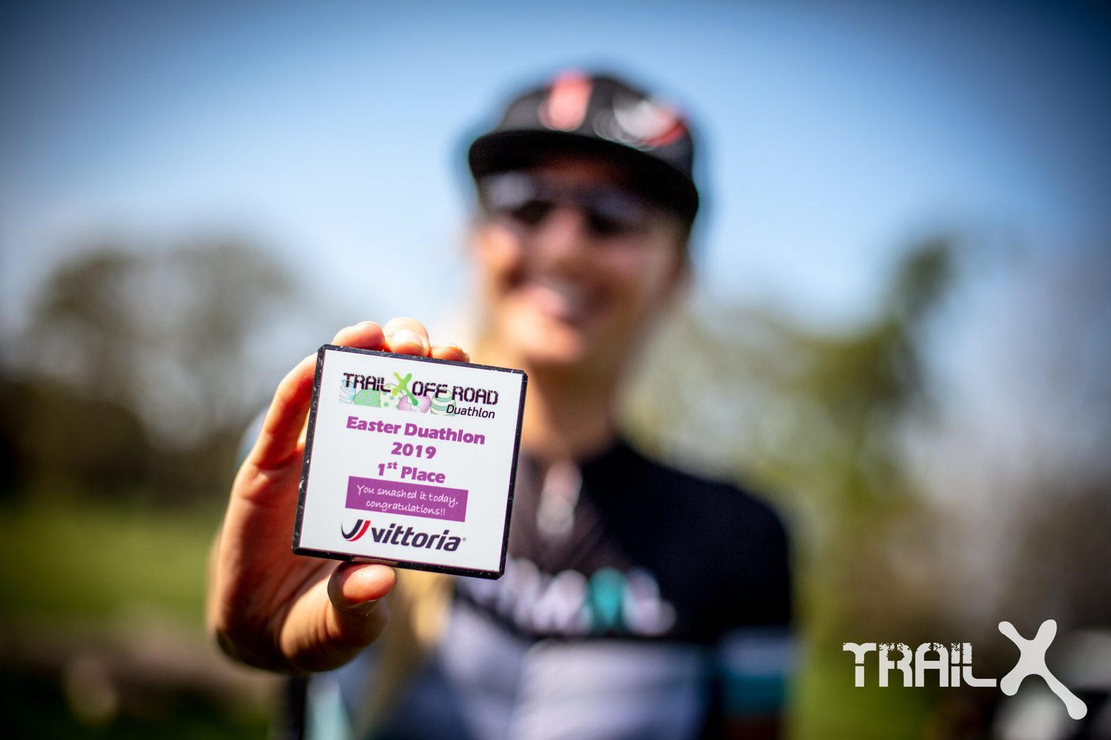 A person in athletic gear, with a blurred background, smiles while holding a small square plaque close to the camera. The plaque reads "TRAIL OFF ROAD Easter Duathlon 2019, 1st Place." The TrailX logo is visible in the bottom right corner of the image.