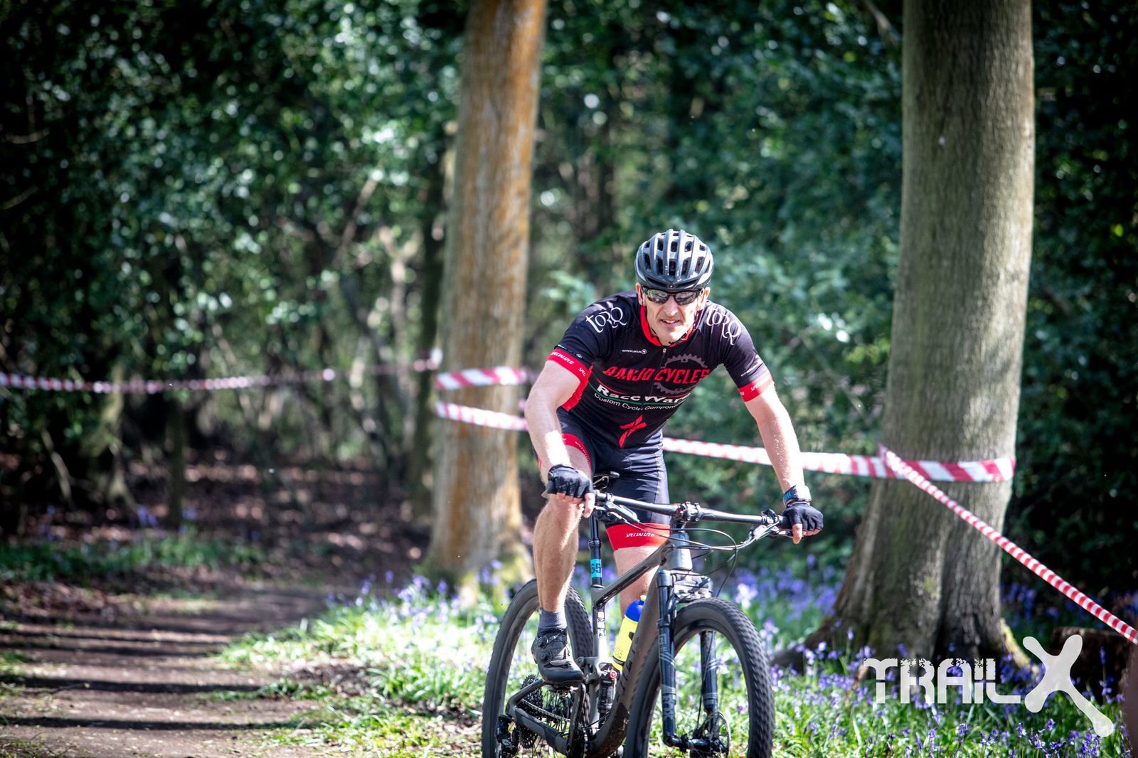 A cyclist in a black and red outfit rides a mountain bike on a forest trail during a race event. The path is lined with red and white tape, and the background is filled with trees and greenery. The cyclist appears focused and determined. The logo "TrailX" is visible.