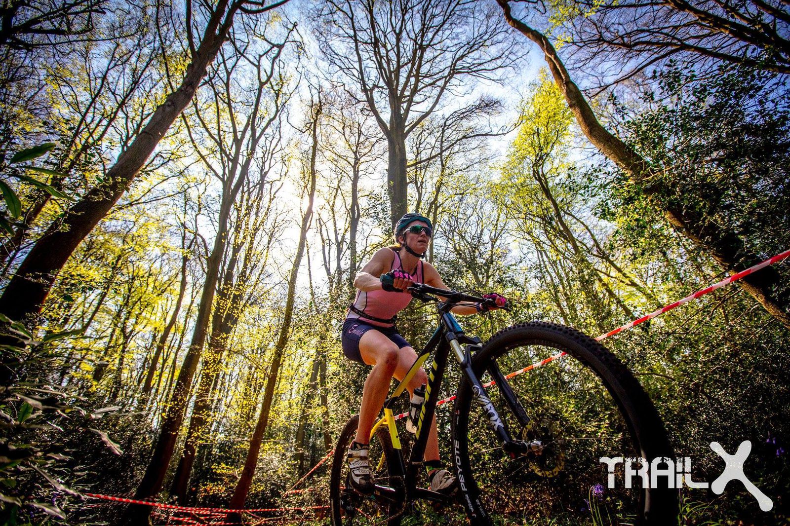 A mountain biker in a pink and black outfit rides aggressively through a forest trail, with sunlight filtering through the trees. A red tape marks the trail, and the cyclist wears sunglasses and a helmet. The "TRI-X" logo is visible in the corner of the image.
