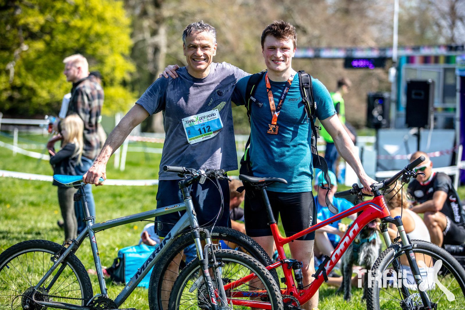 Two men smiling with their arms around each other's shoulders, standing with their mountain bikes in a grassy outdoor area. Both wear athletic gear and race bibs, with one also wearing a medal. In the background, people are sitting or standing near a fenced path and event setup.