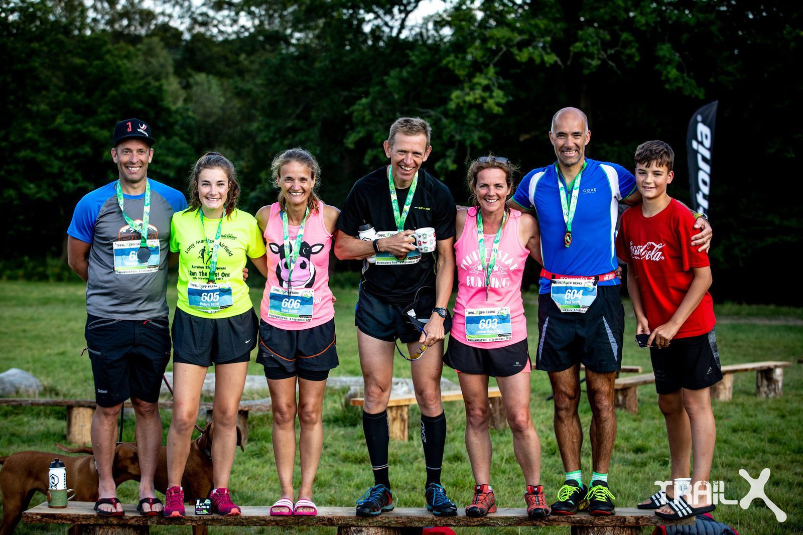 A group of six runners wearing medals poses for a photo outdoors. Four men and two women stand in a line, smiling. The background features trees and grassy terrain. They are dressed in athletic apparel, and one young boy stands on the far right beside the adult group.