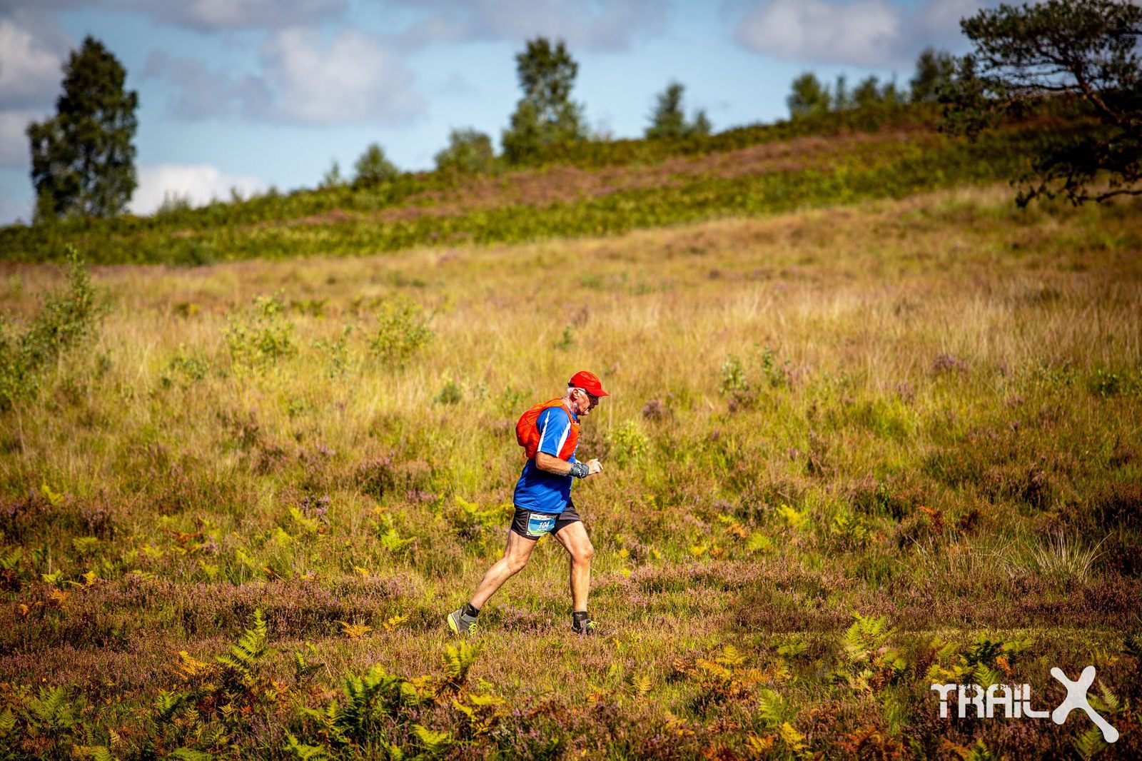 A person wearing a red cap, blue shirt, and shorts is trail running through a scenic grassy landscape with trees in the background. The bright, sunny day highlights the natural surroundings and the person's movement. "TRAIL X" is visible in the bottom right corner.