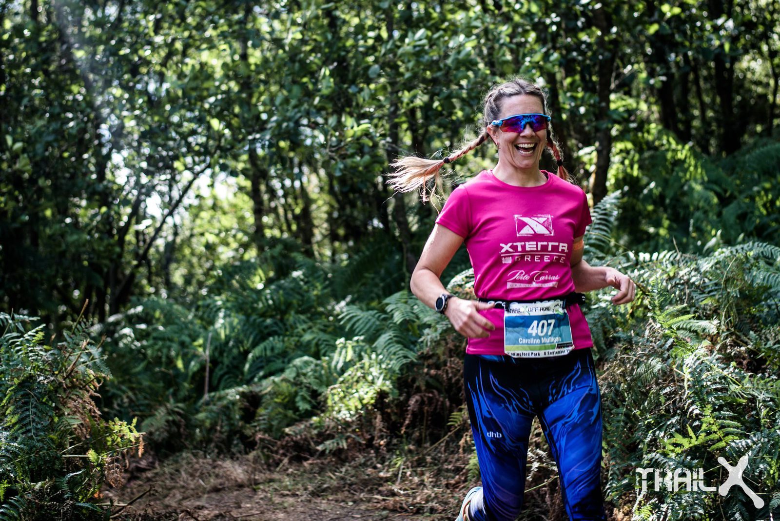 A woman in sunglasses, a pink shirt, and blue athletic pants, with the number 407, smiles as she runs through a lush, green forest path during a race. She appears joyful and energetic amidst the greenery. Trail X logo visible in the bottom right corner.