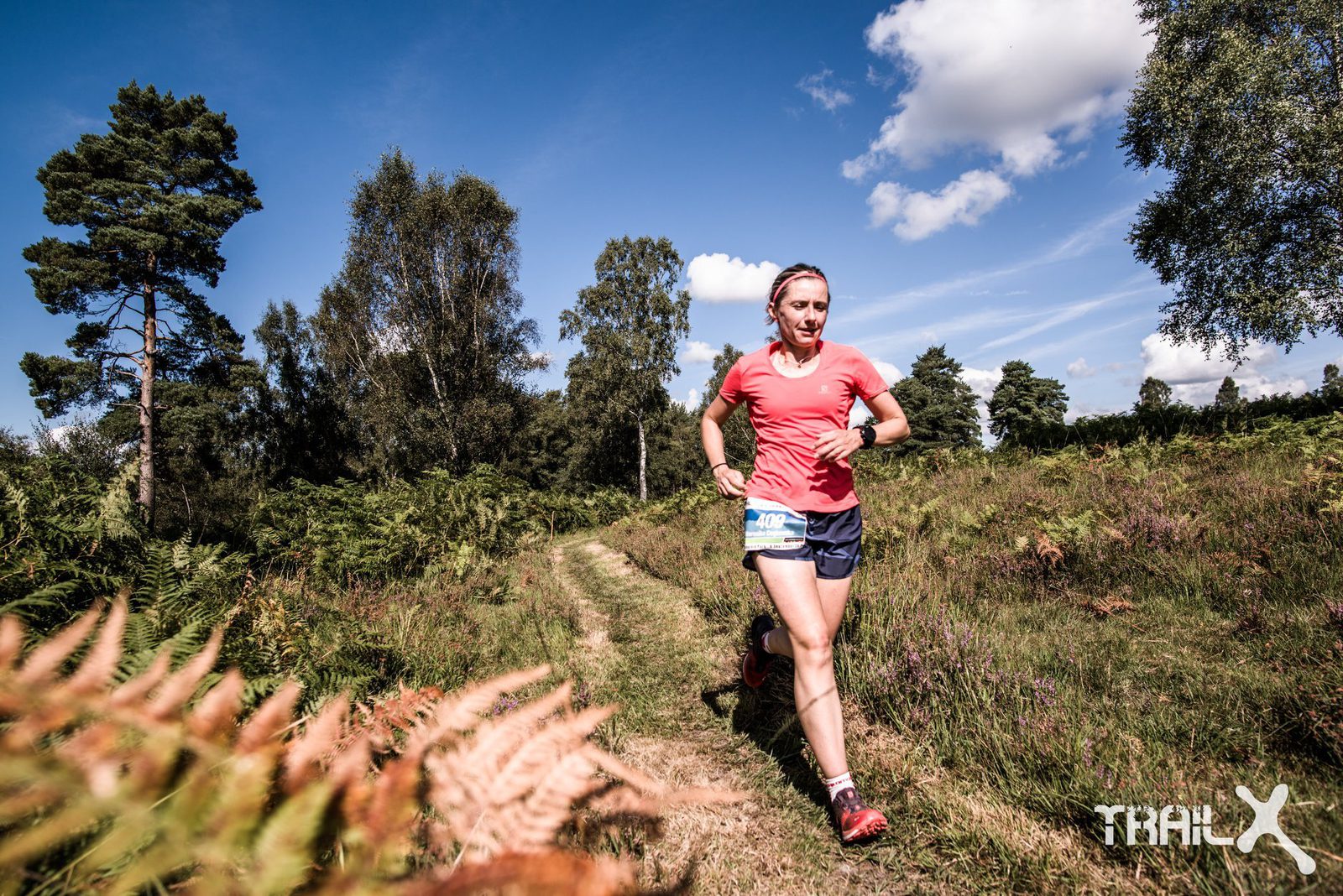 A woman in a red shirt and blue shorts runs on a grassy trail through a lush forest. The sky is clear and blue, with a few scattered clouds. Tall green trees and ferns line the path. The logo "IROMAN" is visible in the bottom right corner.