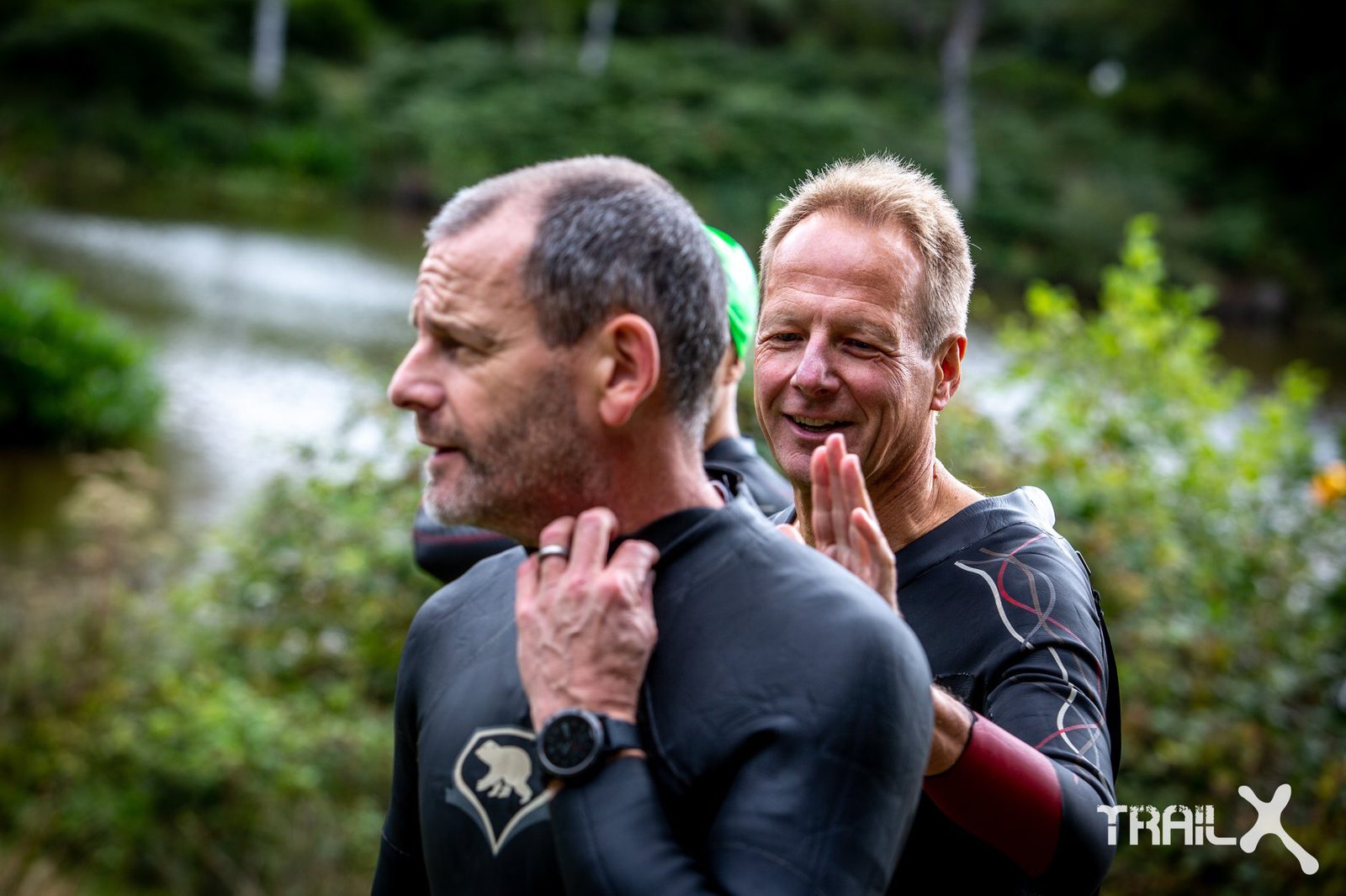Two men prepare for a trail or endurance event beside a body of water. The man in the foreground is adjusting his wetsuit collar, while the man behind him is smiling and patting his shoulder supportively. Both are dressed in dark, athletic wetsuits.