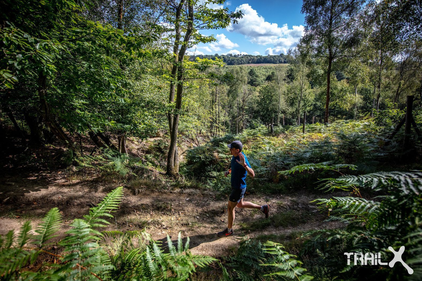A person wearing athletic gear runs on a dirt trail through a lush forest with tall trees and abundant ferns. Sunlight filters through the trees, casting dappled shadows on the path. The sky is partly cloudy, and a "TRAIL X" logo is visible in the lower right corner.
