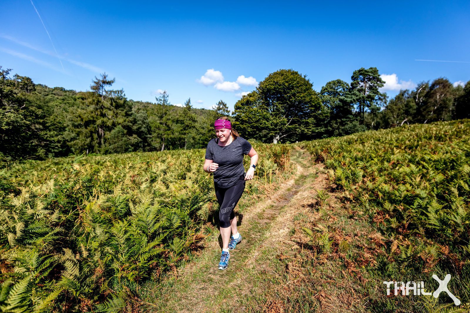 A person wearing a purple headband, black shirt, and black shorts runs on a narrow dirt path through a green, grassy field with trees in the background under a clear blue sky. A race bib is pinned to their leg. The logo "Thail X" is visible in the bottom right corner.