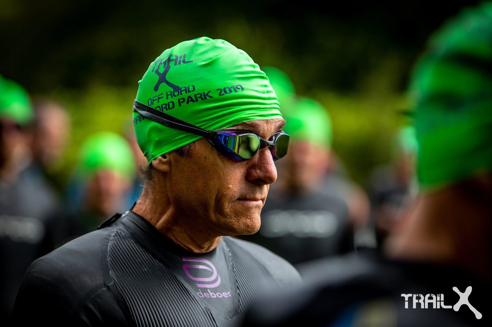 A man in a green swim cap and dark goggles stands in a crowd, preparing for a swim event. He is wearing a black wetsuit, with "Off Road National Park 2019" printed on his swim cap. The background features other participants in similar swim attire. "TRAILX" is branded in the corner.