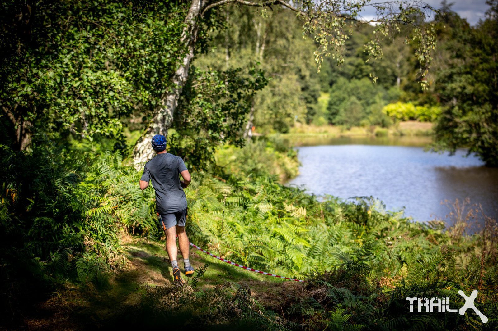 A person in athletic gear runs along a narrow, winding trail beside a calm lake. The path is surrounded by green foliage and trees, with sunlight filtering through. The branding "TRAILx" is visible in the bottom right corner.