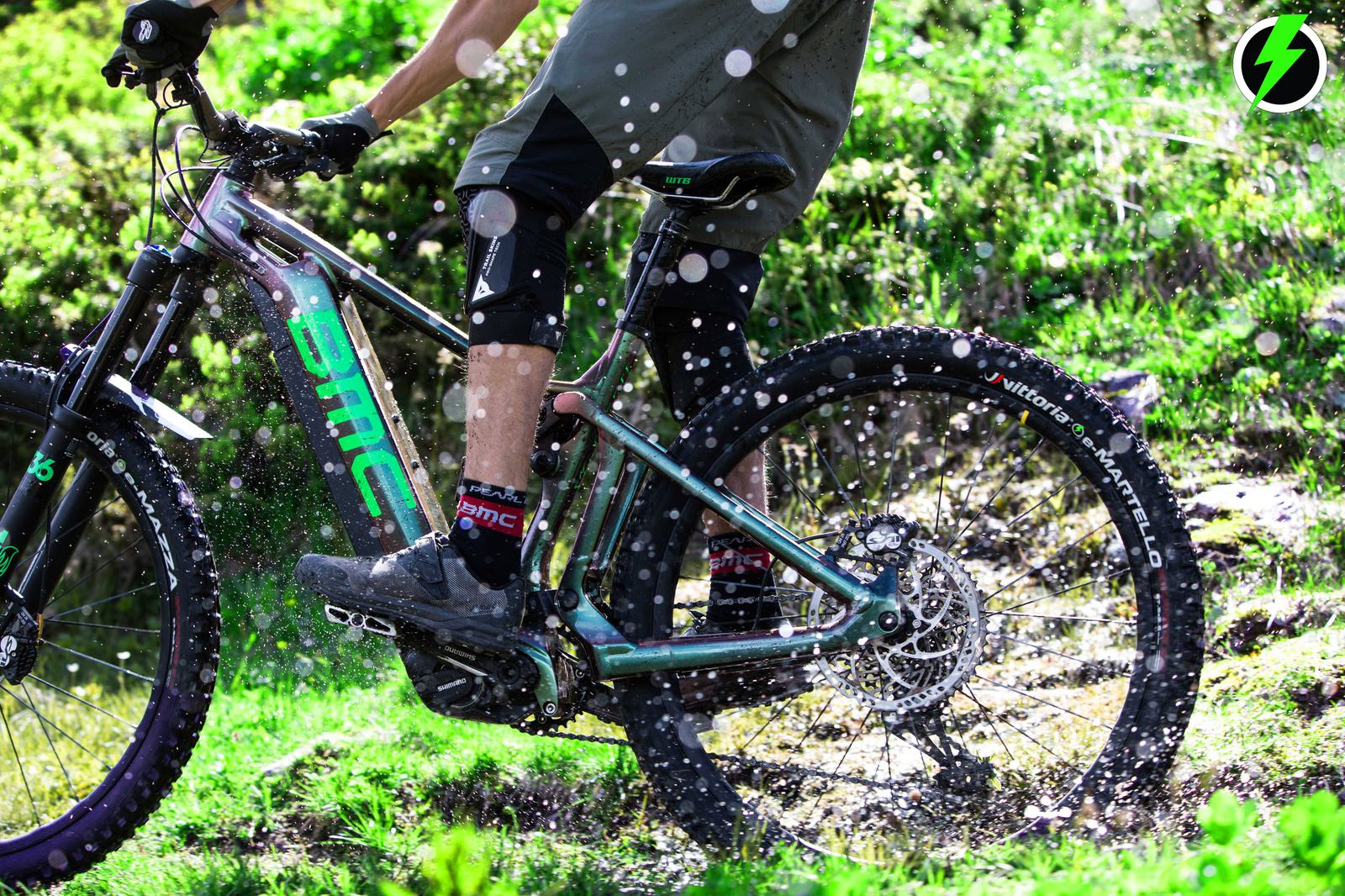 A mountain biker rides a BMC bike through a splash of water on a muddy path surrounded by lush green grass. The rider is wearing knee pads, gray shorts, and black socks with red and white markings.
