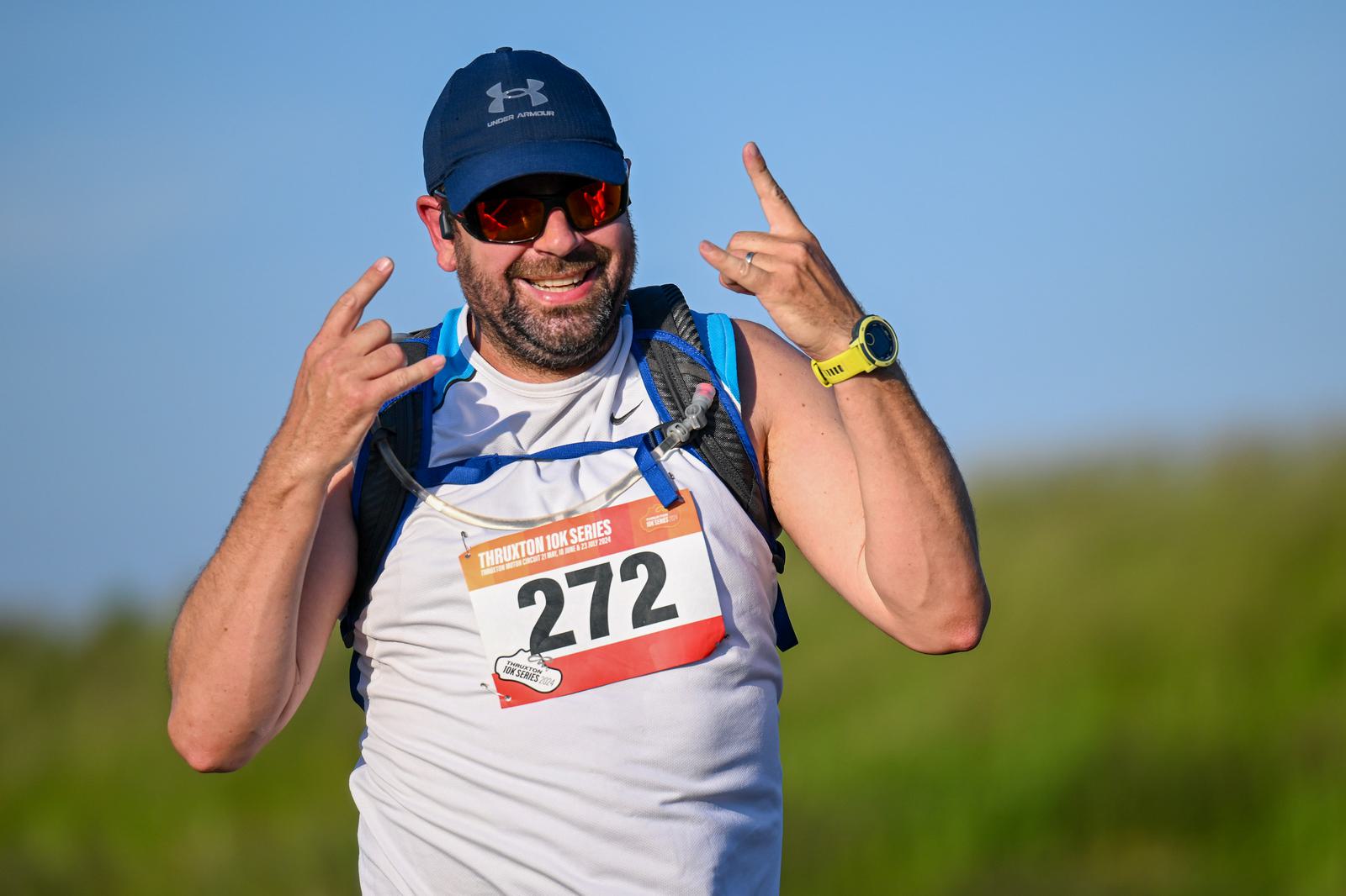 A man in athletic gear, wearing a blue cap and sunglasses, poses with a smile and hand gestures. He's outdoors with a race bib numbered 272 on his chest, against a backdrop of green grass and blue sky.