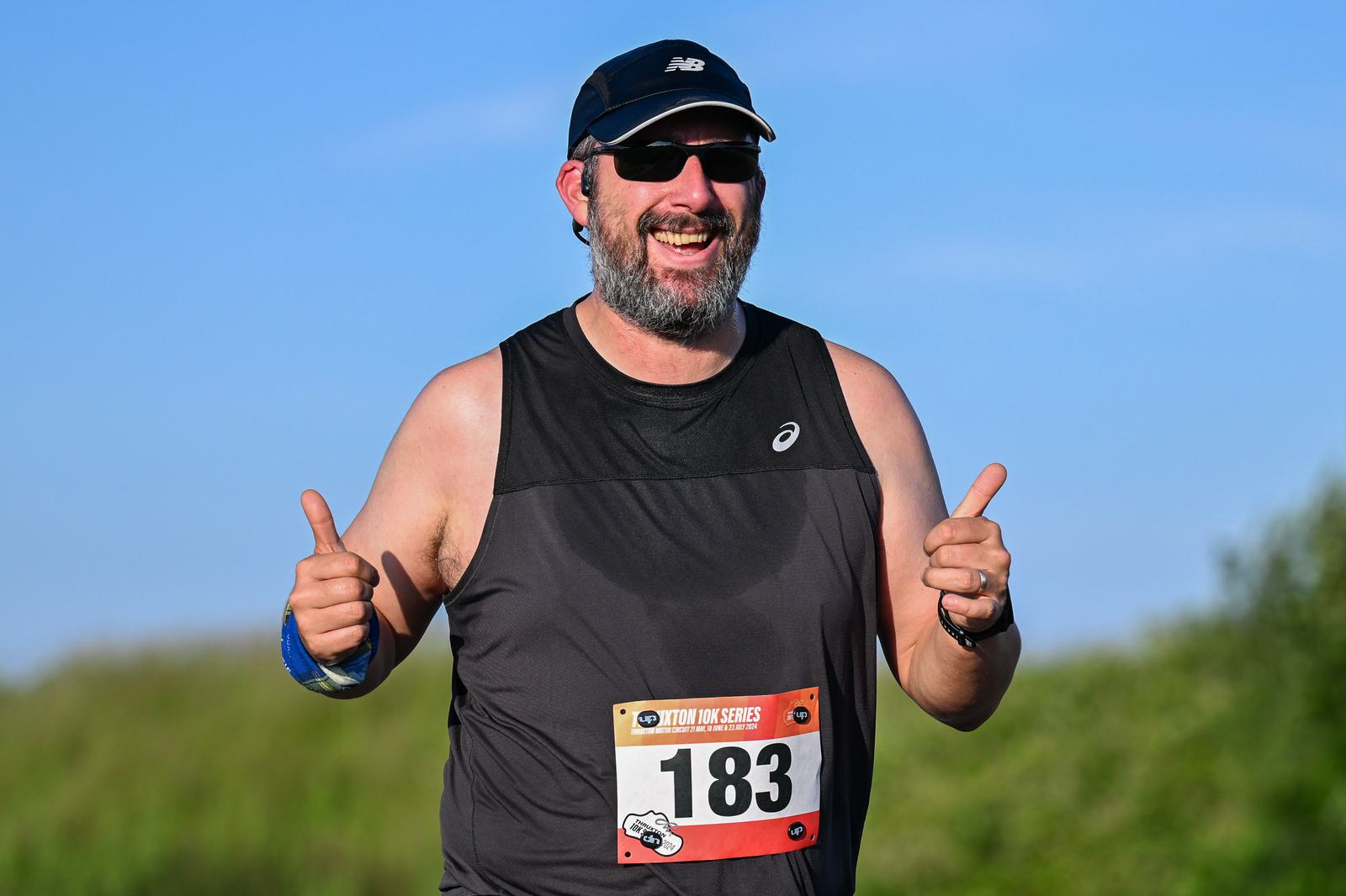 A man wearing sunglasses and a cap is running outdoors, smiling broadly and giving two thumbs up. He's wearing a black sleeveless shirt with a race number 183 pinned on it. The background shows a clear blue sky and green foliage.