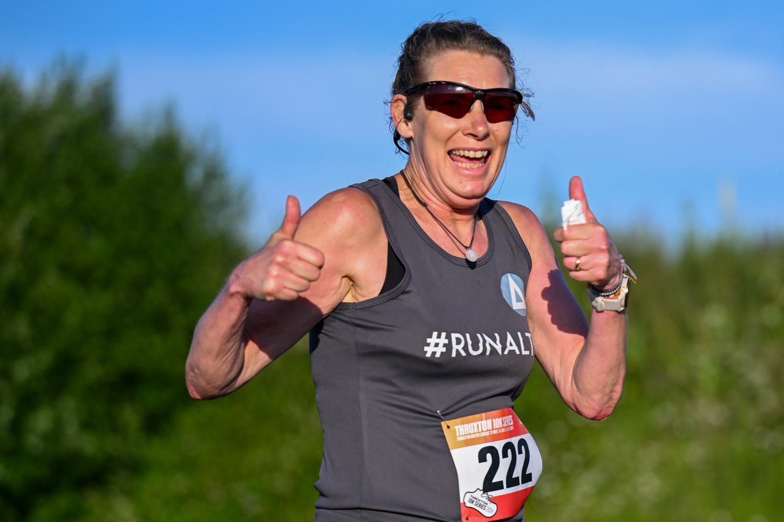 A smiling runner wearing sunglasses and a gray tank top with "#RUNALL" gives two thumbs up while running outdoors. She has a race bib with the number 222. Trees and blue sky are in the background.