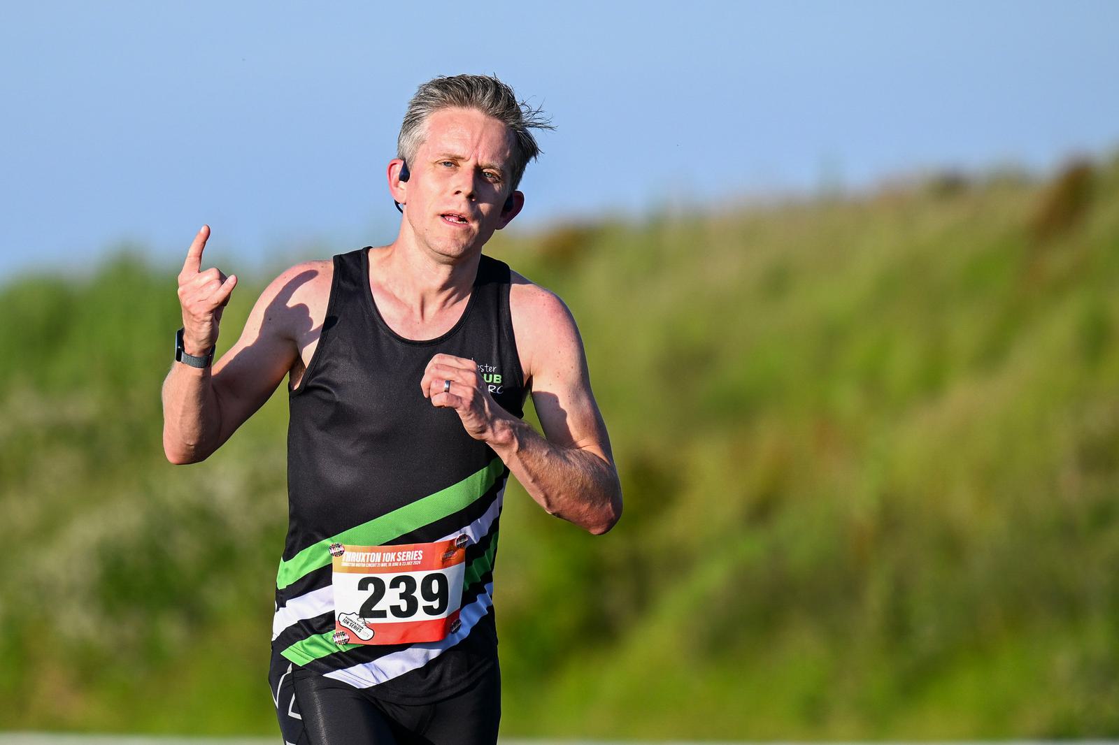 A person with short gray hair is running outdoors, wearing a black tank top with green and white stripes and a race bib number 239. They appear focused and are on a grassy path under a clear blue sky.