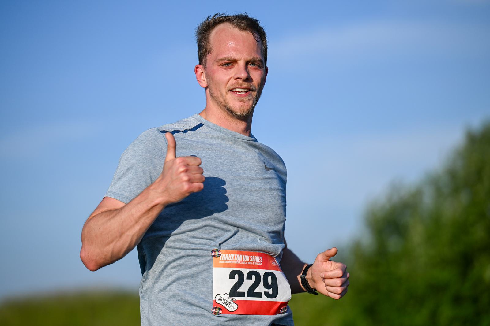 A man in a gray T-shirt is running outdoors with a race bib number 229 on his chest. He is giving a thumbs-up and smiling, with a blue sky and green foliage in the background.