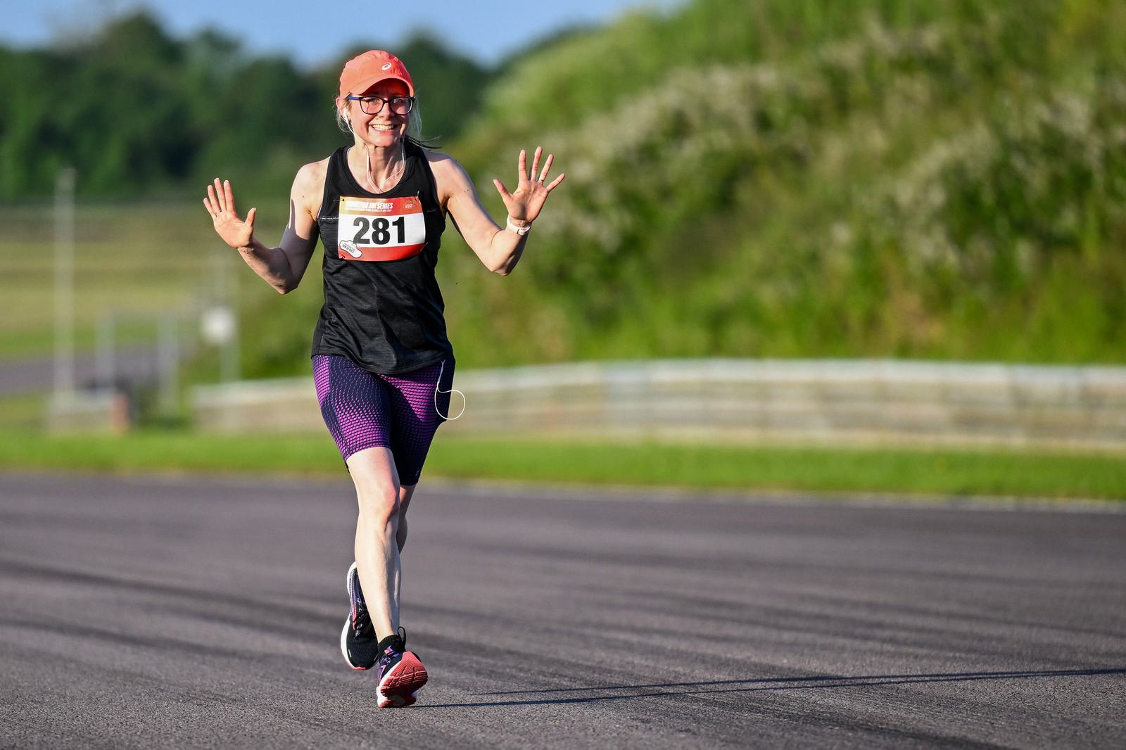 A woman in athletic wear and a cap runs on a track, smiling and raising both hands in the air. She's wearing a race bib with the number 281. The background is grassy with blurry foliage.