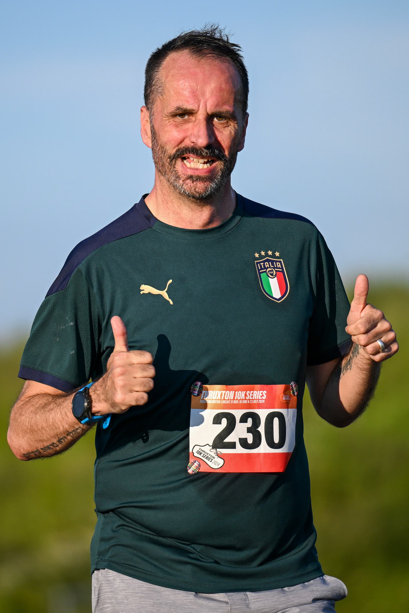 A man outdoors giving two thumbs up, wearing a green sports shirt with an Italian flag emblem and a race bib numbered 230. He has a beard and is smiling, with a clear sky and greenery in the background.
