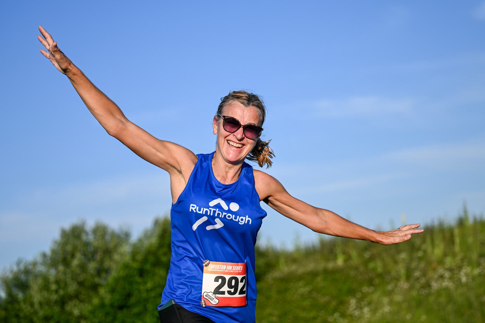 A woman in sunglasses and a blue "RunThrough" tank top joyfully extends her arms while running outdoors. She wears a race bib numbered 292. The background features a clear blue sky and greenery.