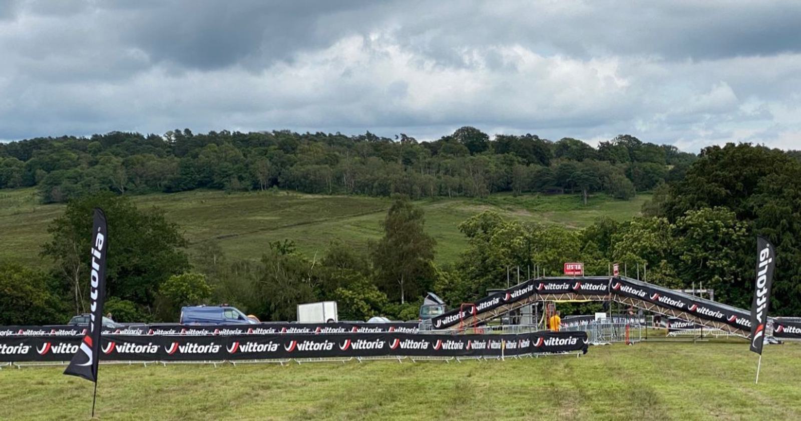 A grassy field set up for a sporting event, bordered by banners with a "Vittoria" logo. There are a few vehicles and equipment in the background, with lush green hills and trees under a cloudy sky.