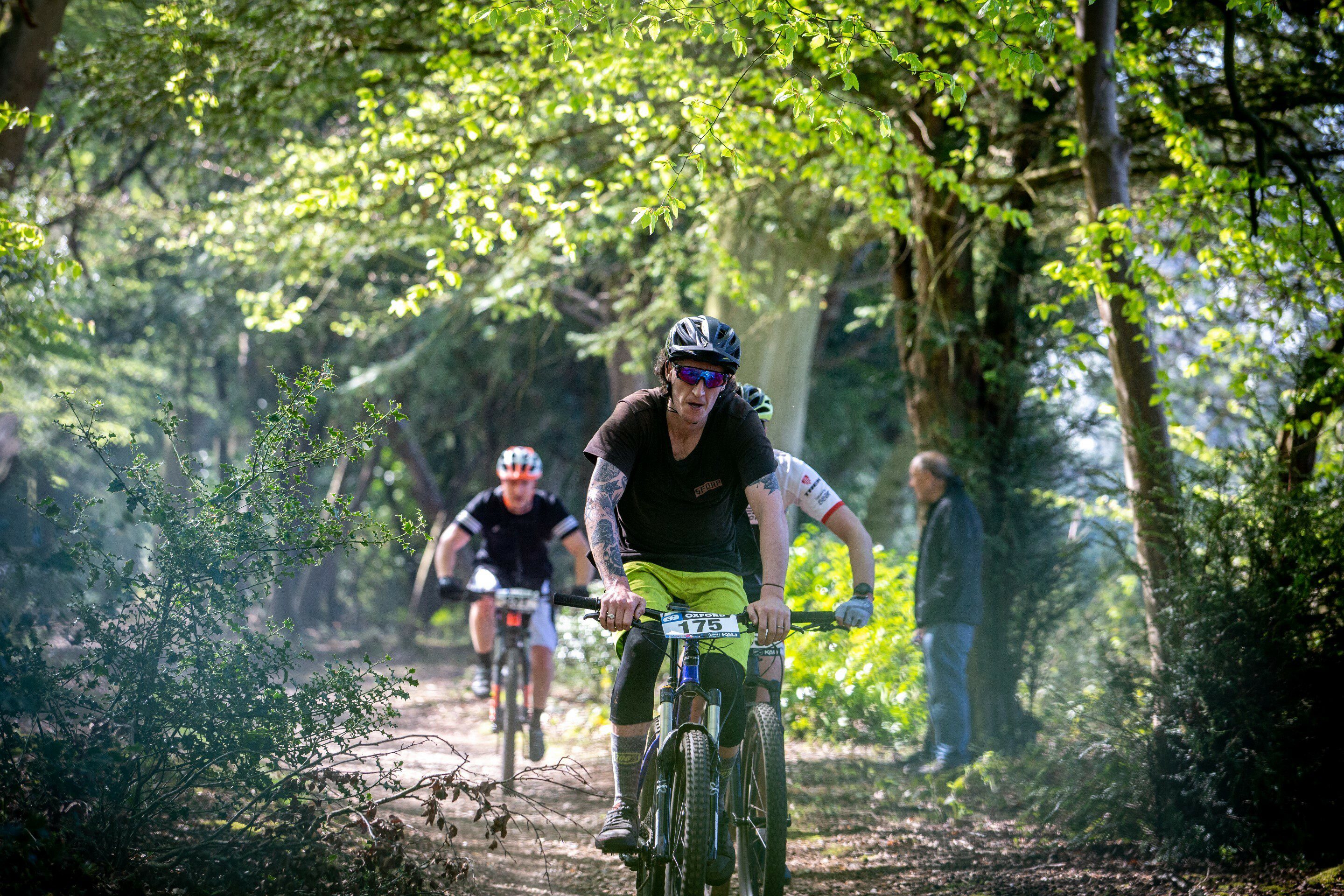 Three cyclists ride their bikes on a dirt trail through a forest, with one cyclist in the foreground wearing bright yellow shorts and sunglasses. Sunlight filters through the trees, casting a dappled light on the path. A person stands to the side observing.