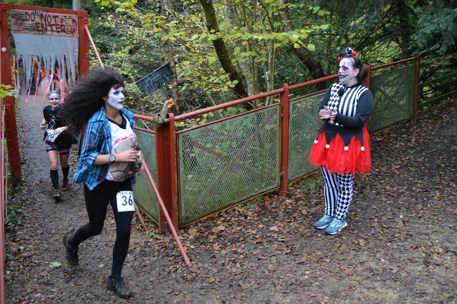 A person with face paint and long hair runs through a wooded area holding a doll and wearing the number 36. Behind them is a person in a black and red harlequin outfit and face paint, standing near a gate with a sign that says "Do Not Enter.