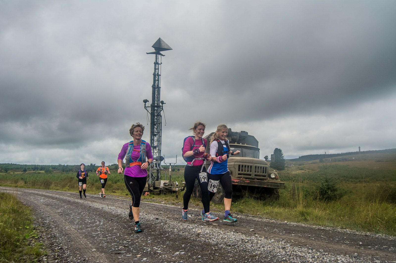 A group of runners, including three women in colorful athletic gear, are jogging on a gravel path during an outdoor race. Behind them is a vintage military truck and a tall radar tower. The sky is overcast, adding a dramatic backdrop to the rural landscape.