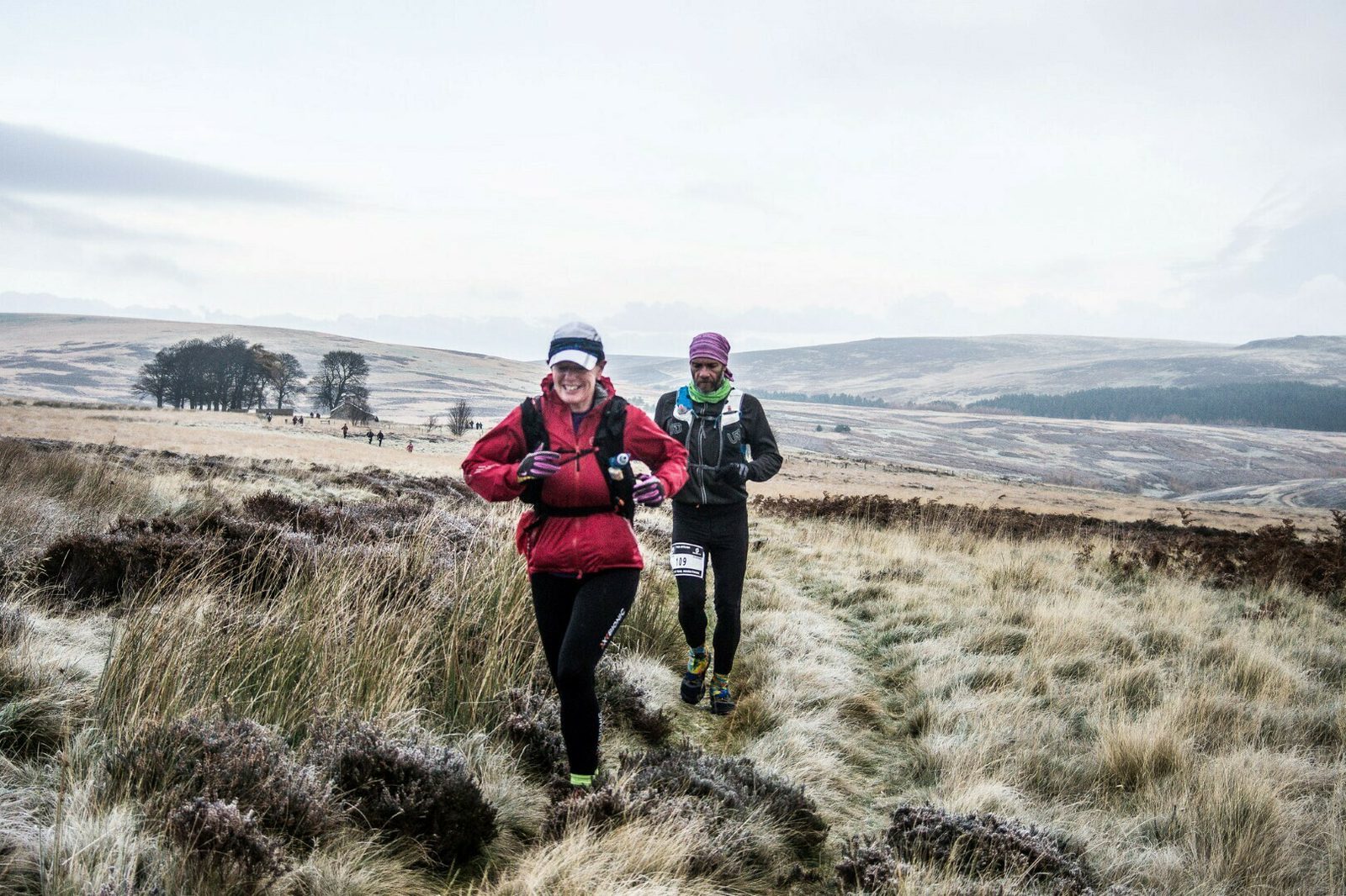 Two runners are on a trail through a grassy, open moorland. The leading runner, wearing a red jacket and black leggings, is smiling, while the second runner, wearing a blue jacket and purple hat, follows. The landscape is vast with gentle hills and sparse trees.