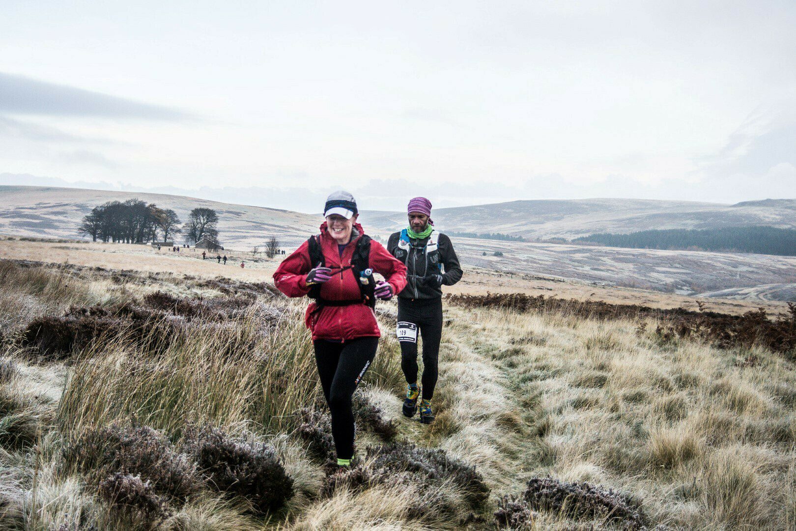 Two runners are on a trail through a grassy, open moorland. The leading runner, wearing a red jacket and black leggings, is smiling, while the second runner, wearing a blue jacket and purple hat, follows. The landscape is vast with gentle hills and sparse trees.
