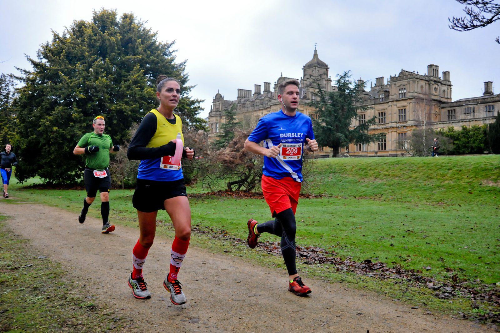 A group of runners participate in a race on a dirt path with a large, historic building in the background. The participants are wearing race bibs and sports attire, with trees and greenery surrounding the path. The sky is overcast.
