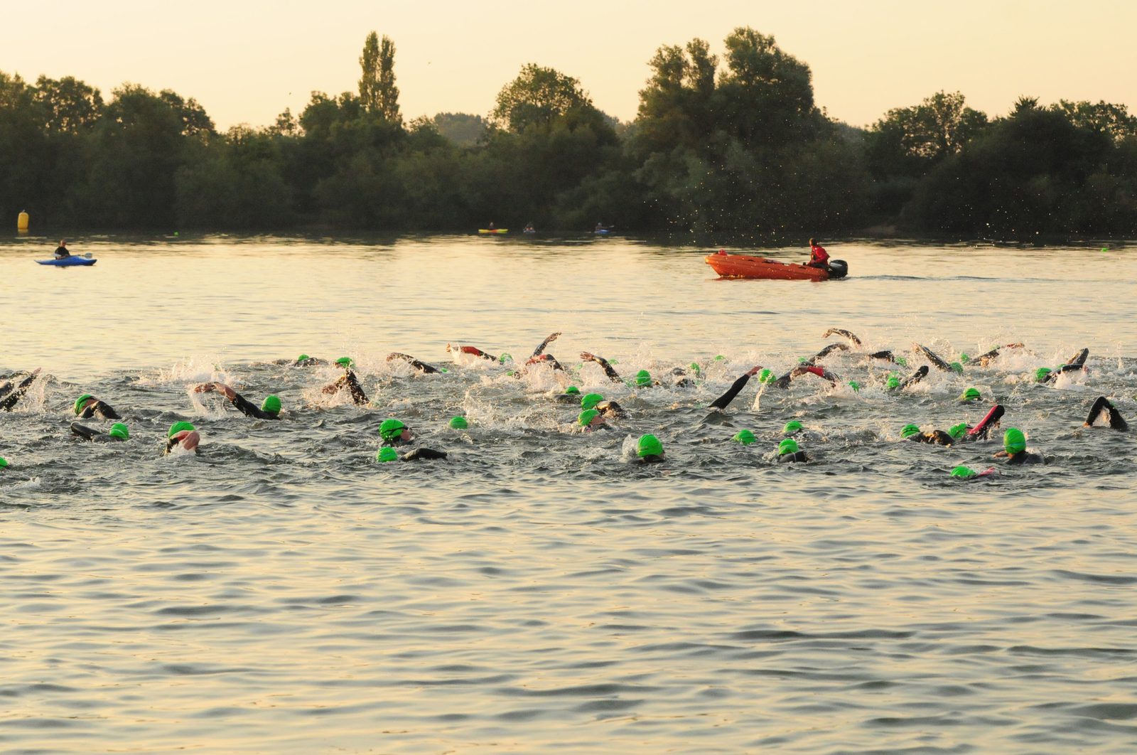Swimmers wearing green swim caps compete in an open water race at sunrise. The swimmers are dispersed across the water while a person in an orange kayak supervises nearby. The background features lush trees and a calm lake.