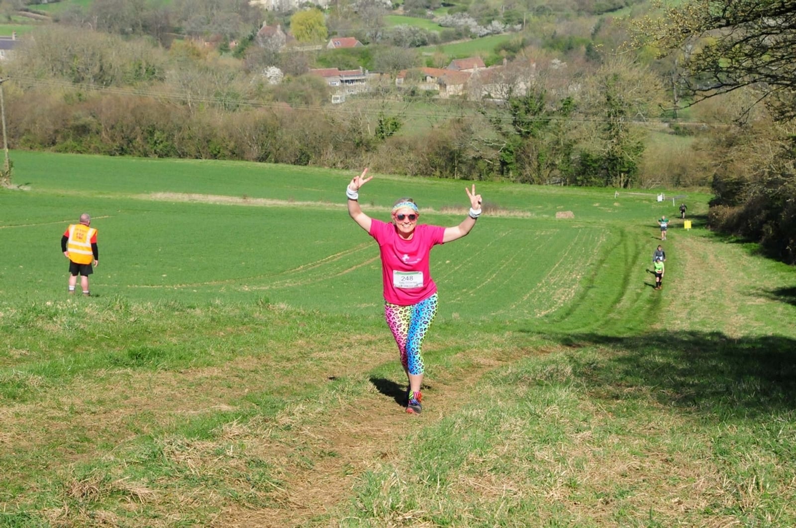 A runner in colorful workout attire, including a pink shirt and rainbow-patterned leggings, runs through a grassy field during a race. The runner is smiling and showing a peace sign with both hands. Other runners and a race official are visible in the background.