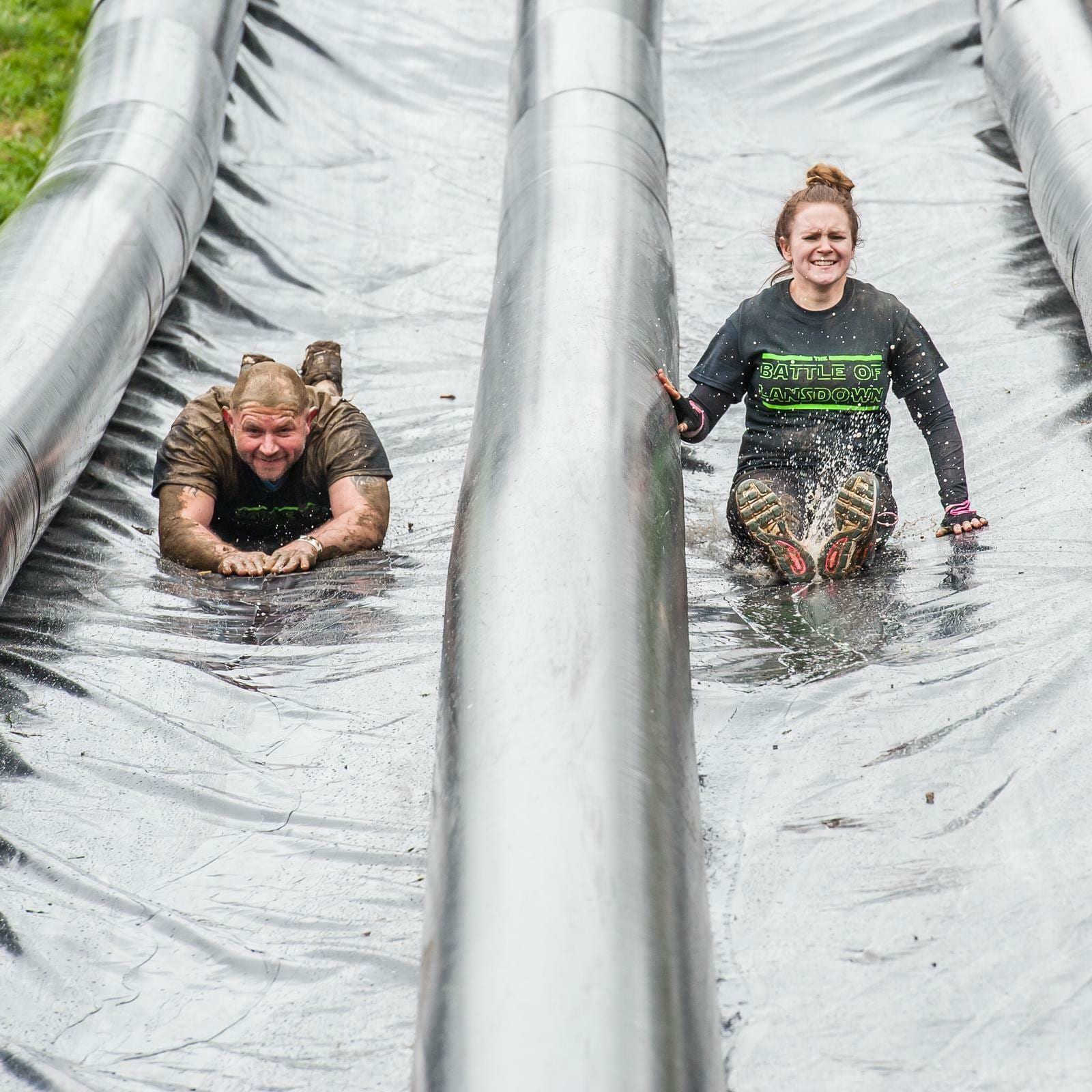 Two people sliding down parallel inflatable water slides outdoors. The person on the left is sliding face down and covered in mud, while the person on the right is sliding seated and smiling. They appear to be participating in an obstacle course event.