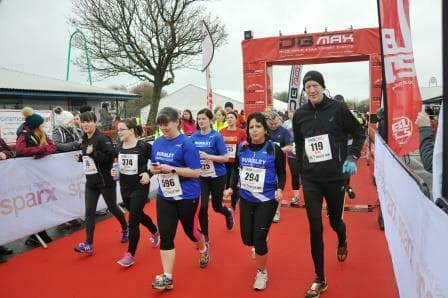 A group of runners, including women and men, cross the finish line at a race event. They are wearing race bibs and athletic gear. The finish line is marked by a red archway with banners. Other people and buildings are visible in the background.