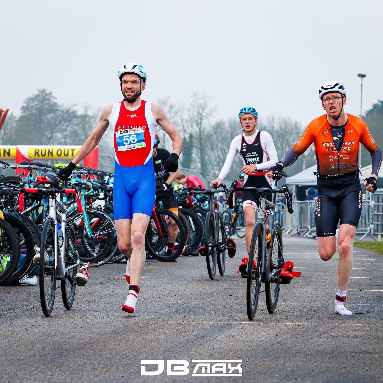 Three triathletes in gear are running with their bikes through a transition area. They wear helmets and numbered vests. Behind them, other bikes are racked. The background shows a "RUN OUT" sign. The area is overcast and appears cool.