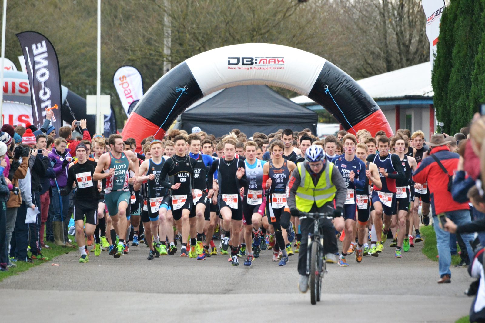 A group of athletes is running at the start of a race, following a cyclist who leads the way. They are under a large inflatable arch with event branding. Spectators are gathered on the sides.