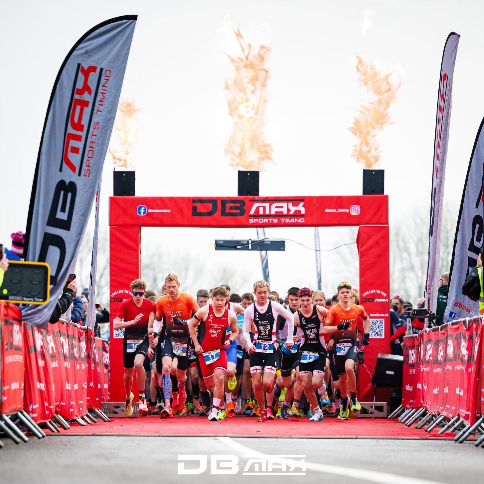 Group of athletes start a race at the DB Max Sports Timing event. They are running under an archway with flames above. A crowd watches from behind barriers, with two flags on either side displaying the event logo.
