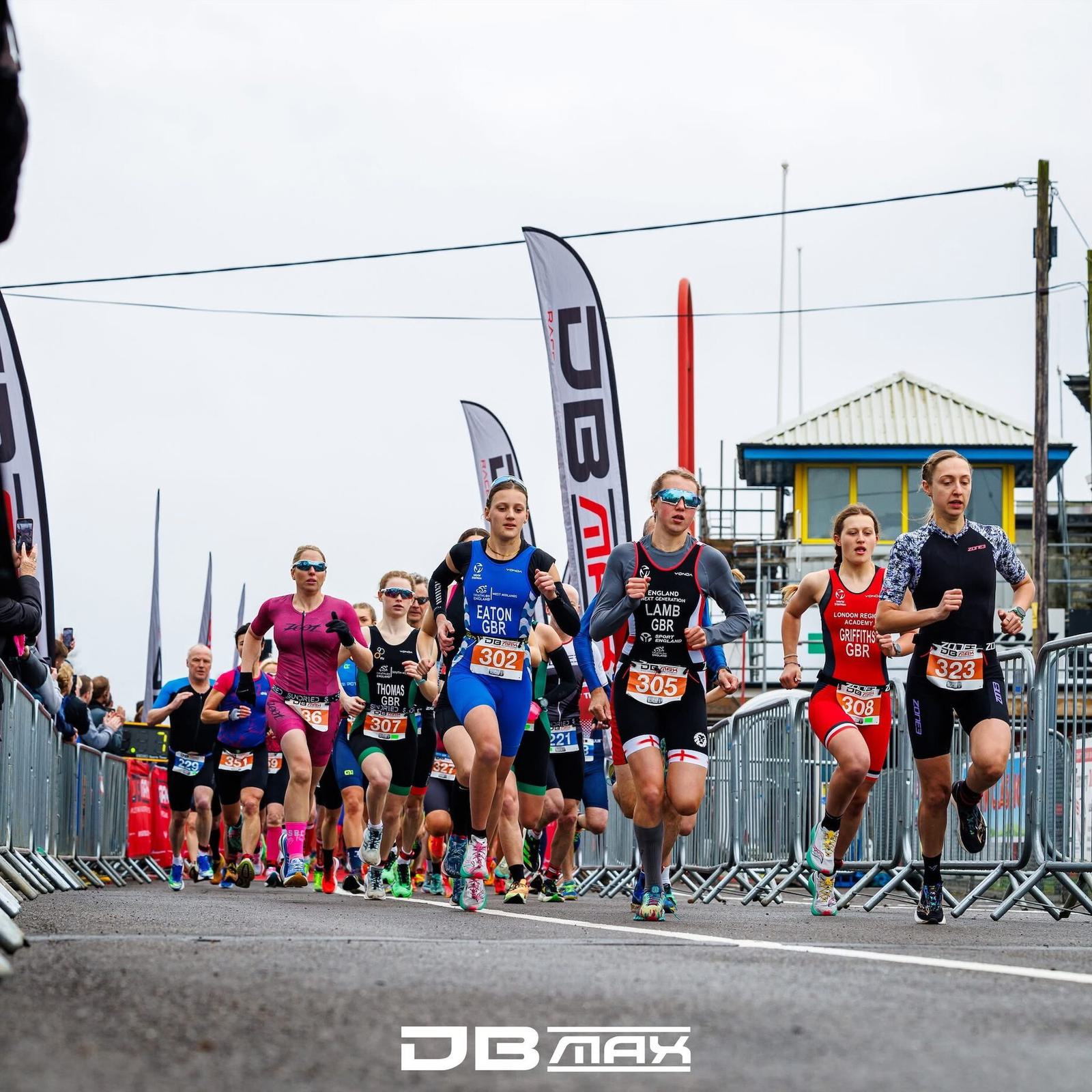 A group of athletes in colorful sportswear competes in a road race. The runners are surrounded by metal barriers and DB Max flags, with a cloudy sky overhead. Each participant wears a unique race bib with numbers.